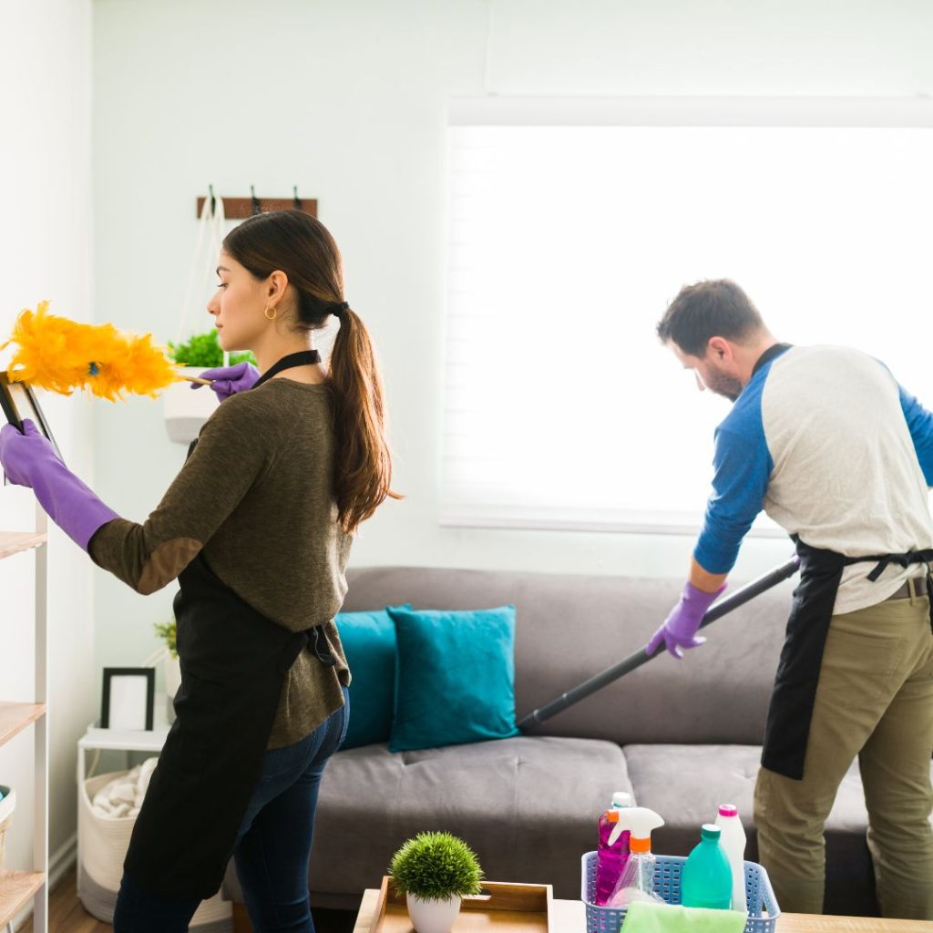 A woman dusting a picture frame and a man is cleaning the couch